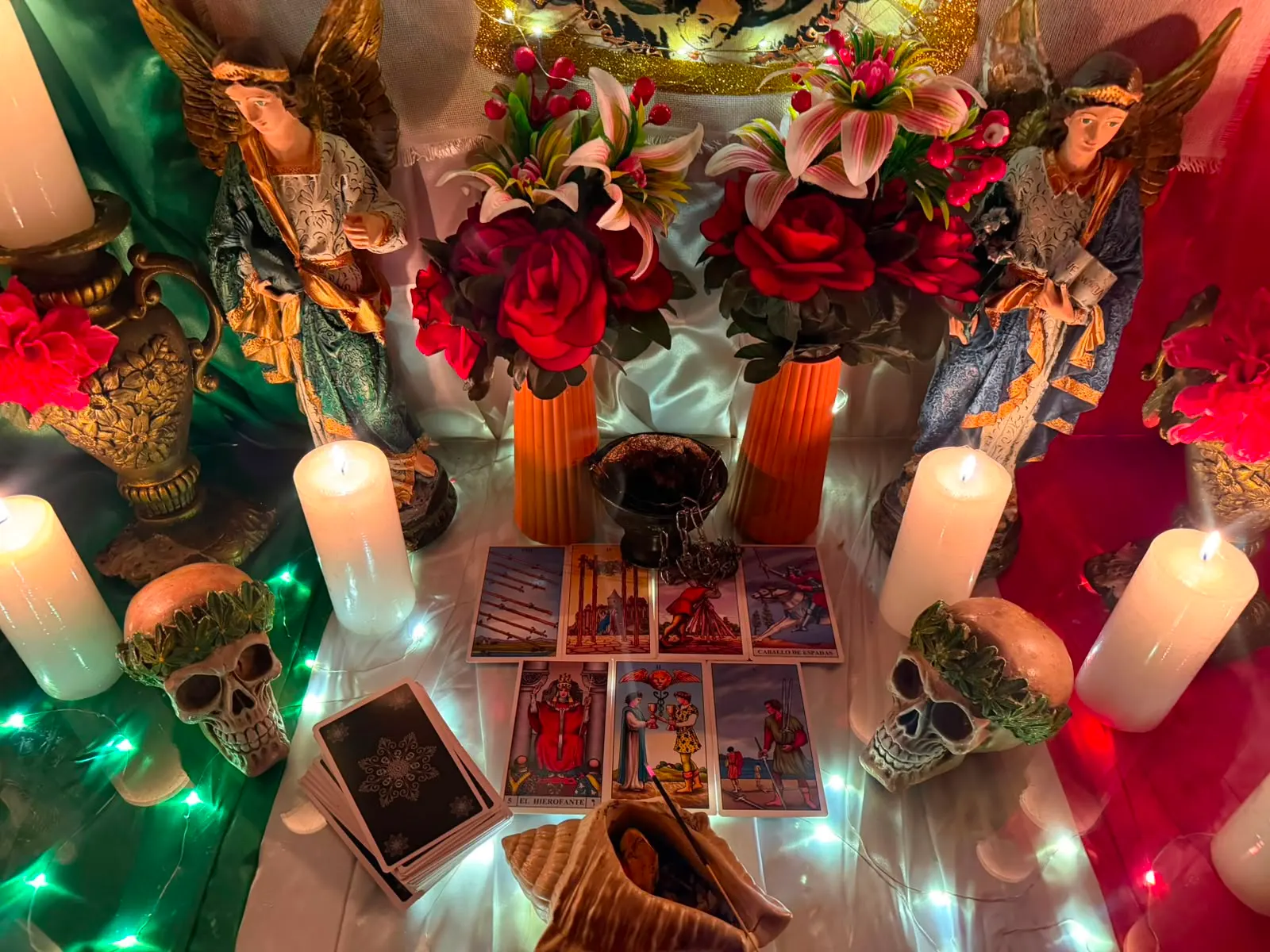 Altar con calaveras decoradas, ángeles, flores rojas, velas encendidas y cartas sobre el mantel